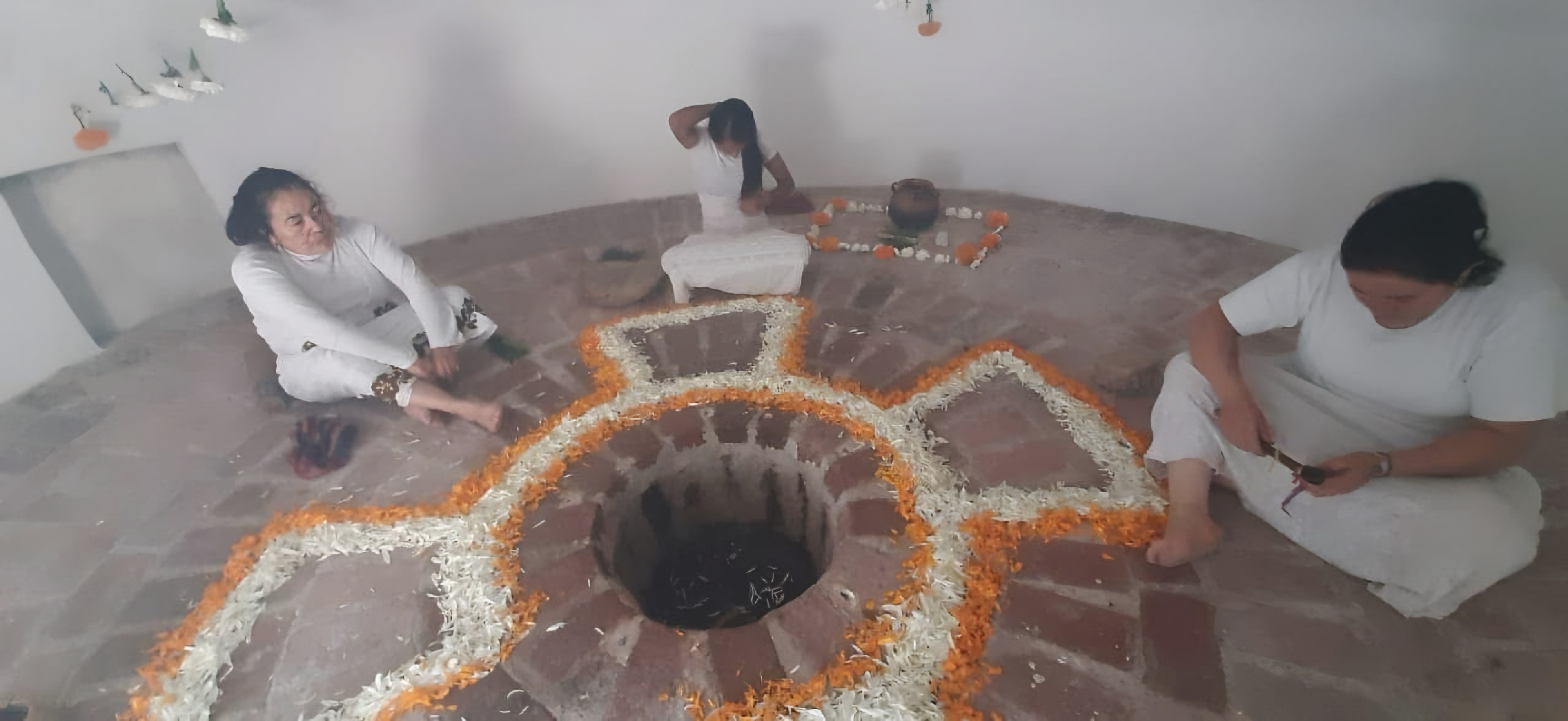 Mandala de pétalos naranjas y blancos alrededor del pozo de piedras, con tres mujeres vestidas de blanco