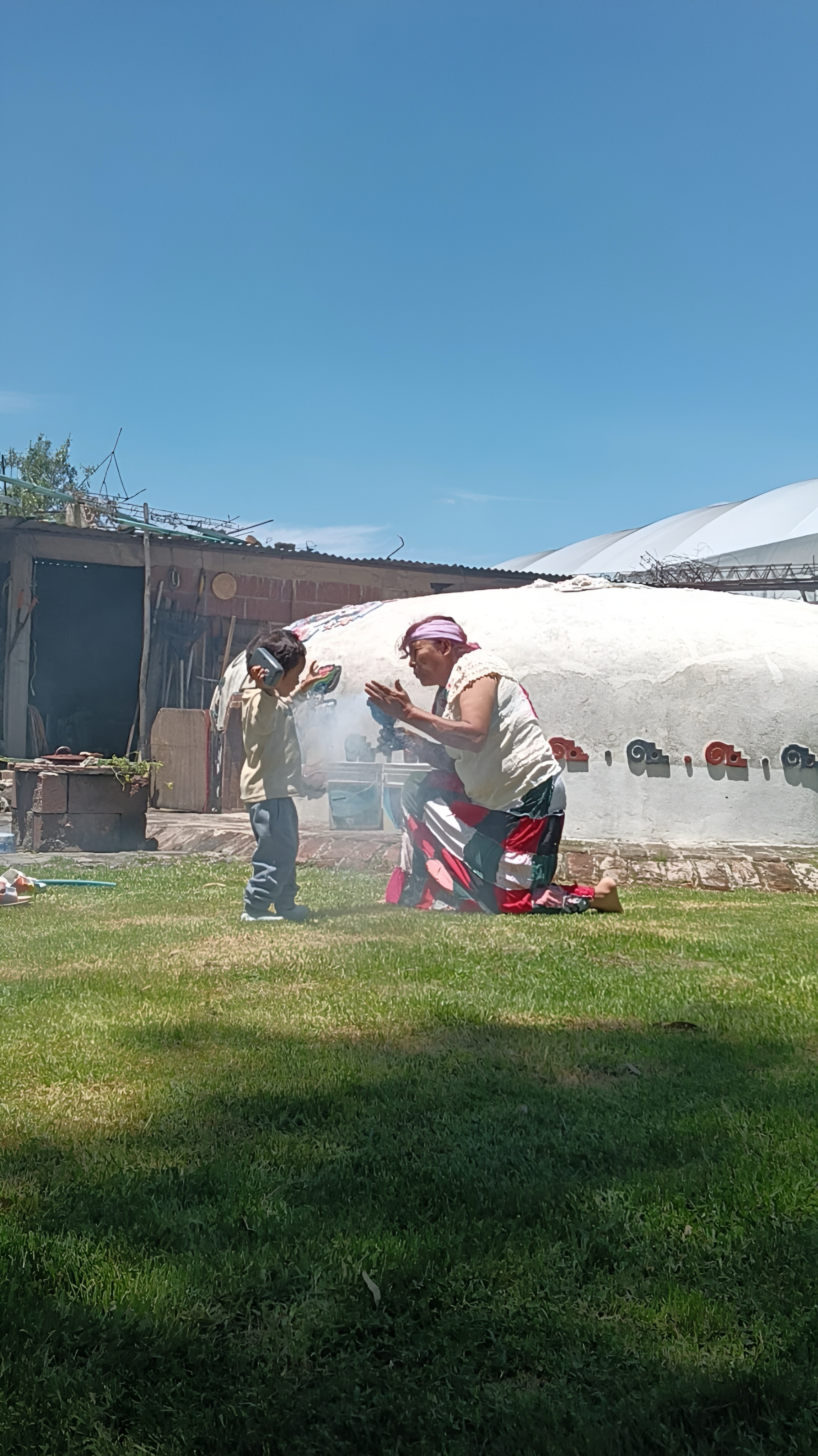 Una abuela arrodillada sahúma con copal a un niño frente al temazcal